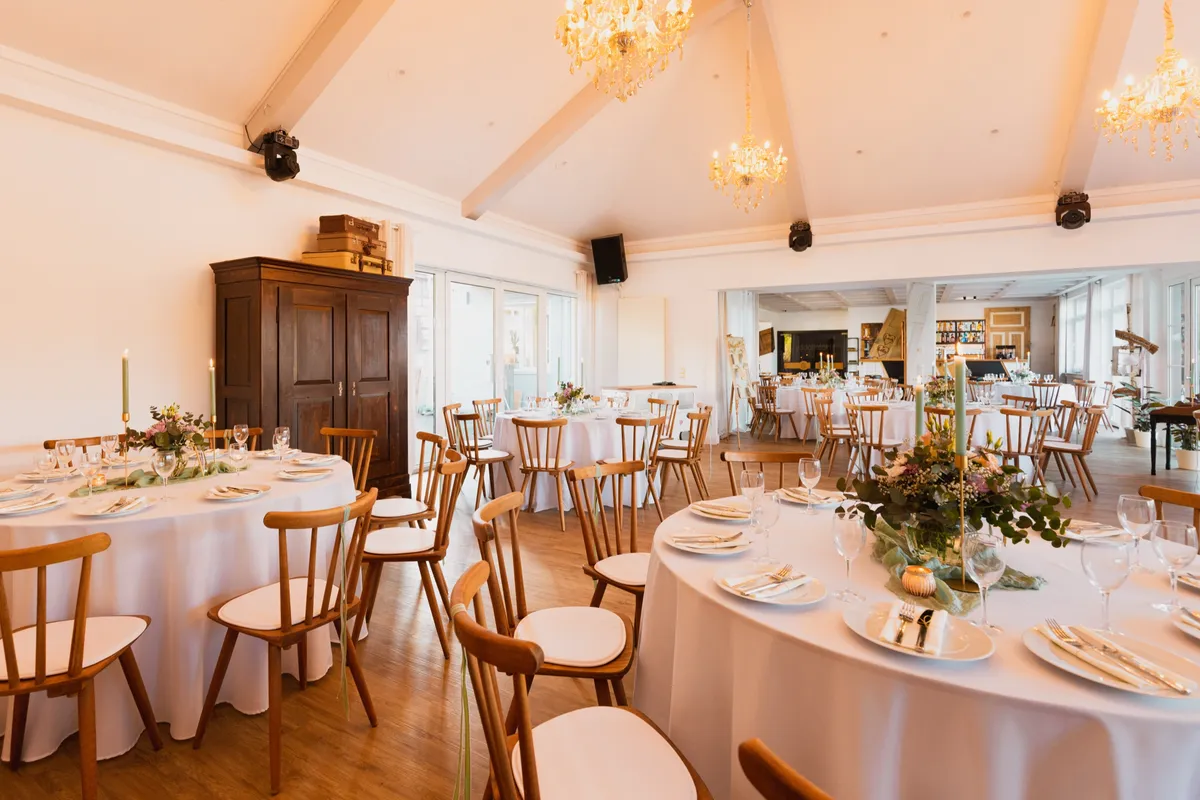 Heller Festsaal mit Kronleuchtern, Gewölbedecke und rund eingedeckten Hochzeitstafeln