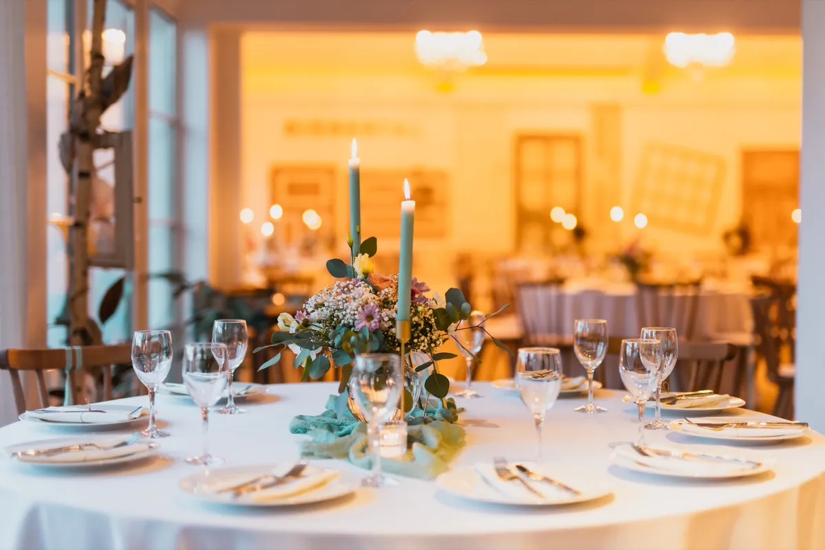 Eingedeckter runder Hochzeitstisch mit Blumen und Kerzen, dahinter der beleuchtete Festsaal