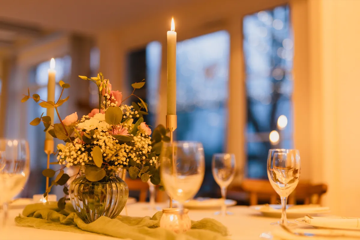Tischdekoration mit Blumengesteck, brennenden Kerzen und Weingläsern zur blauen Stunde im Festsaal